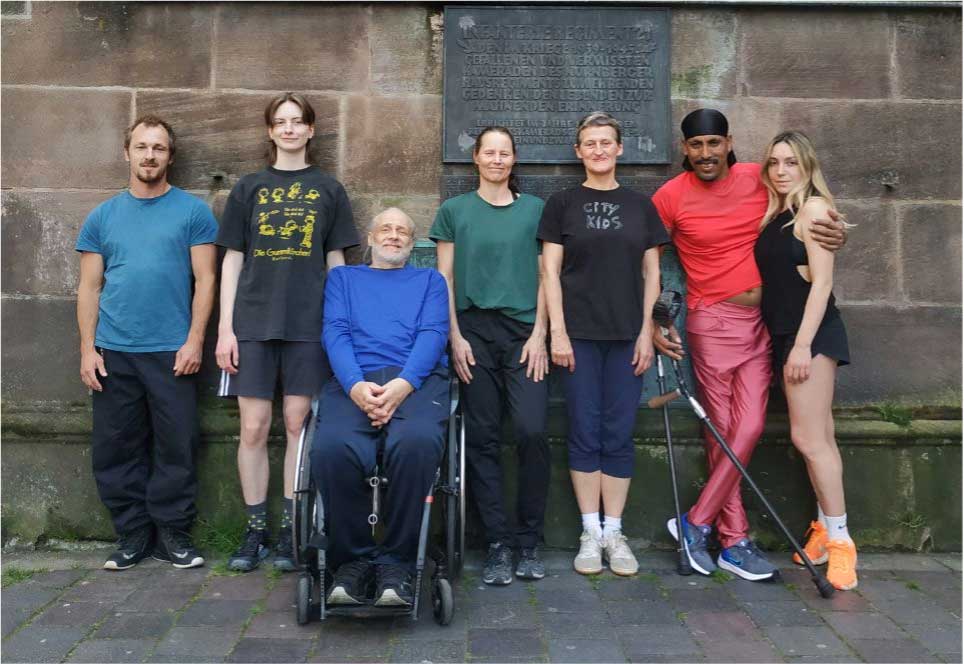 Die 7 Personen von der Tanz·gruppe Every·Body sind nebeneinander vor einer Mauer.