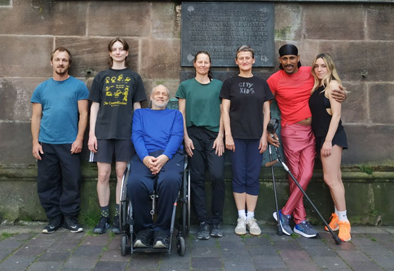 Die 7 Personen von der Tanz·gruppe Every·Body stehen nebeneinander vor einer Mauer.